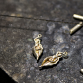 Bild på två silverhängen från Goddess-kollektionen under tillverkning på en arbetsbänk. Hängena visar en kvinnofigur med pärla i snäckform, med öglor för halsband. Verktyg syns runt omkring.