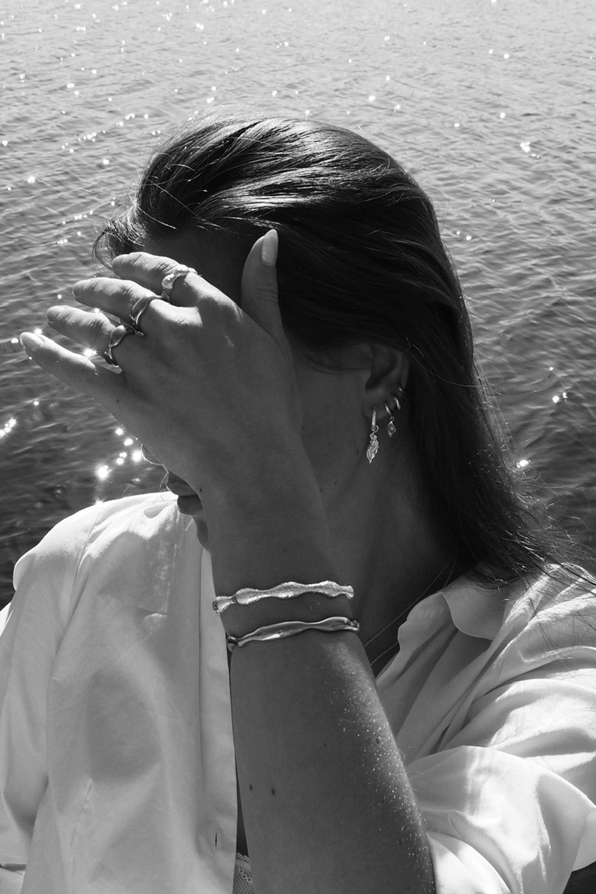 Svartvit bild på en kvinna i brunt hår vid havet. Bärandes olika smycken från OYS jewelry. Solen skiner. 