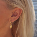 Bild på ett örhänge från Goddess-kollektionen, visat på modell. Örhänget är en liten guldtonad ring med ett hängande hänge som föreställer en kvinnofigur med pärla i snäckform.